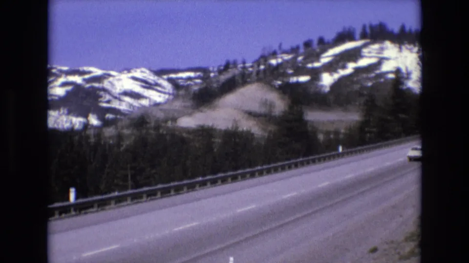 A mountain highway curves past snowy wooded slopes under a blue sky, with a small portion of a passing vehicle visible at the far right edge.