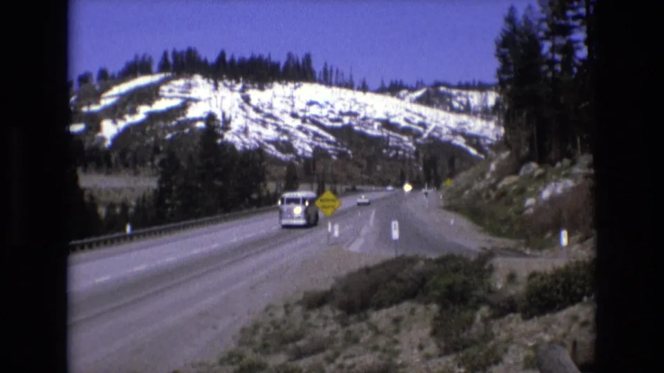 1973 8mm frame of traffic on a mountain highway, including a light-colored van, smaller cars, a yellow roadside warning sign, guardrails, and snow-streaked hills.