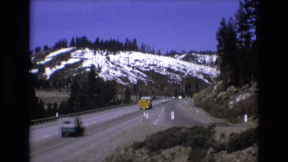 A car passes in the foreground on a mountain highway while distant vehicles, yellow warning signs, evergreen trees, and snow-dusted slopes fill the background.