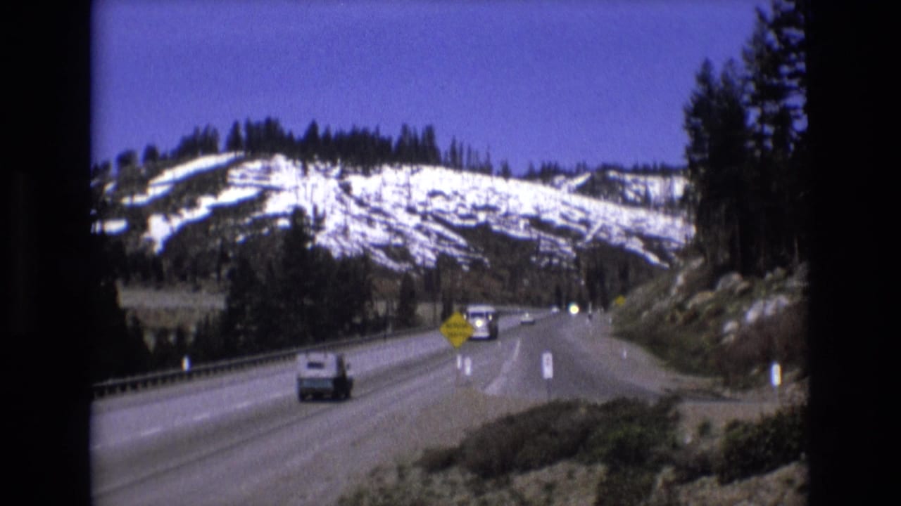 Mountain highway with cars, snow-dusted hills, and evergreen trees under a blue sky.