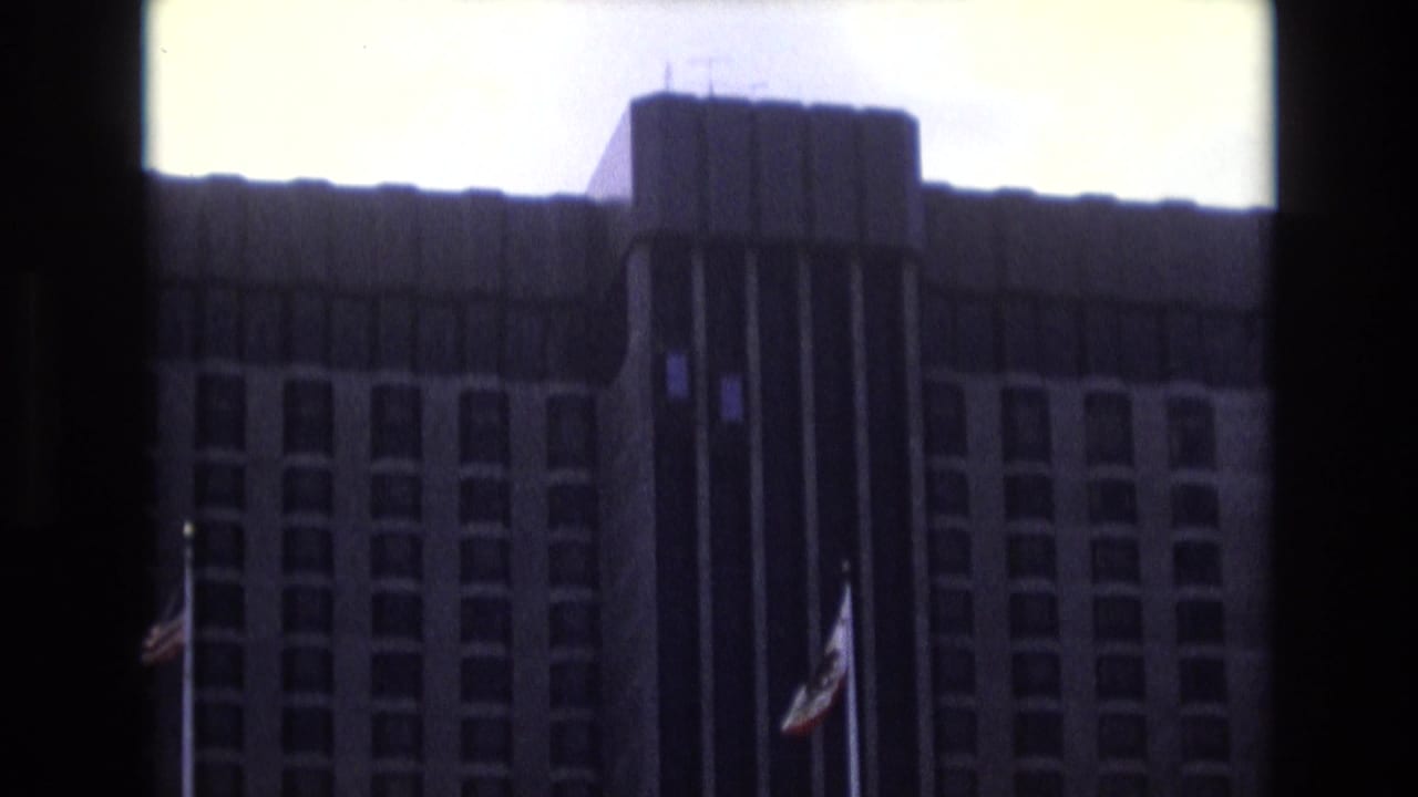 1973 8mm travel footage frame showing a large high-rise building exterior with rows of windows, a dark central vertical section, and flags on poles in front.