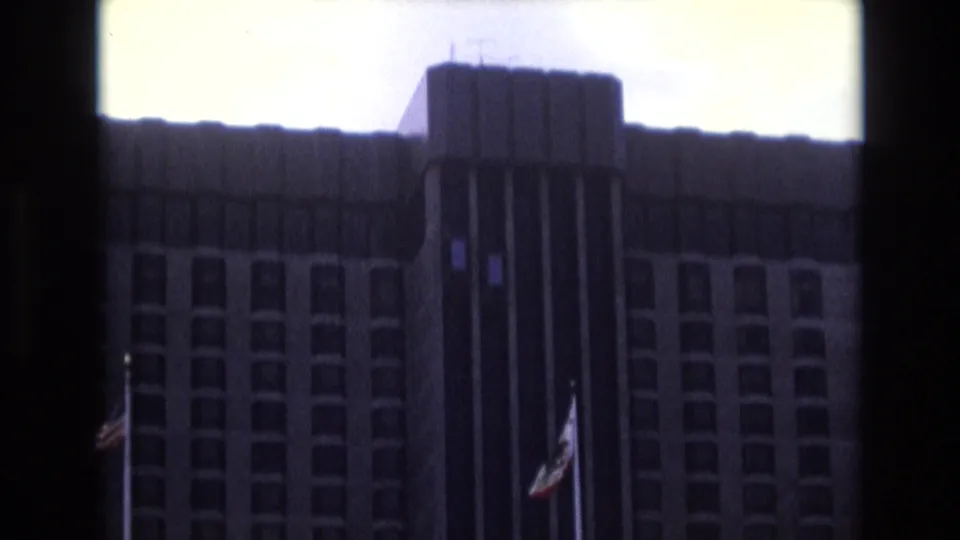 1973 8mm travel footage frame showing a large high-rise building exterior with rows of windows, a dark central vertical section, and flags on poles in front.