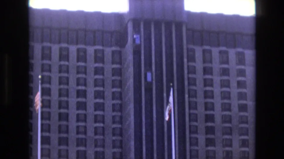 Soft 1973 8mm frame of a large building exterior with repetitive window bays, a dark central strip, and flagpoles carrying American and California flags.