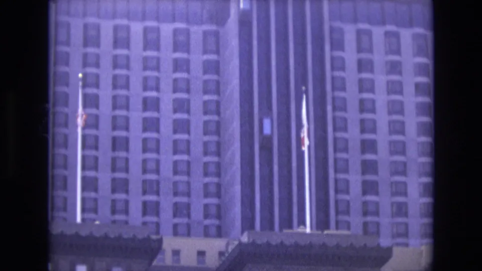 1973 8mm travel footage frame showing a closer view of the high-rise exterior, with rows of windows, vertical architectural lines, and American and California flags near the entrance area.