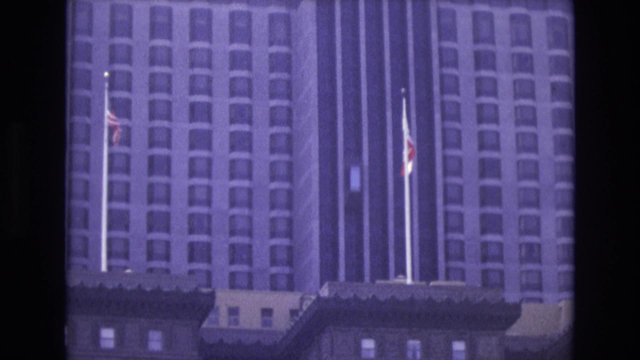 Tall city building with flags above darker foreground rooftops.