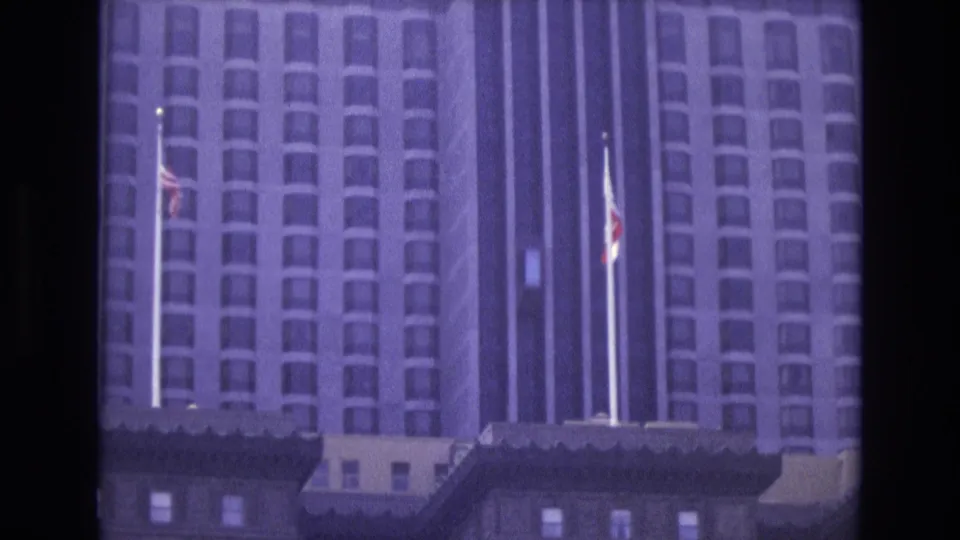 1973 San Francisco 8mm frame showing a tall hotel-like facade, flagpoles, and darker older buildings in the foreground.
