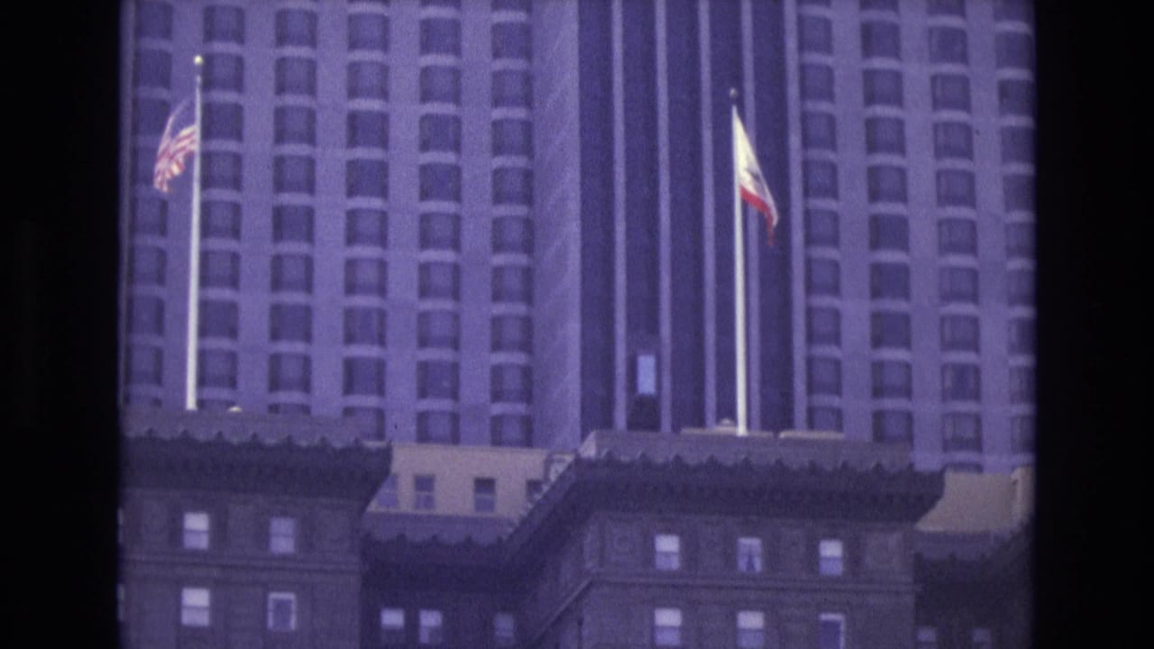 1973 San Francisco frame with American and California flags rising in front of a tall downtown building facade.