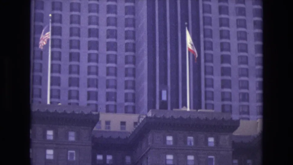 1973 San Francisco frame with American and California flags rising in front of a tall downtown building facade.