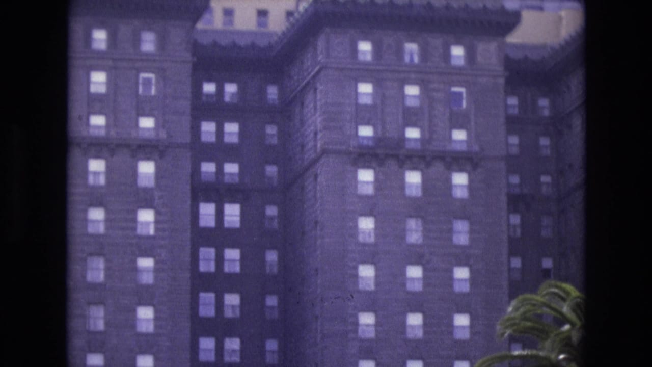 1973 San Francisco 8mm frame of a historic hotel exterior with repeated window rows, decorative upper trim, and palm fronds entering from the right.