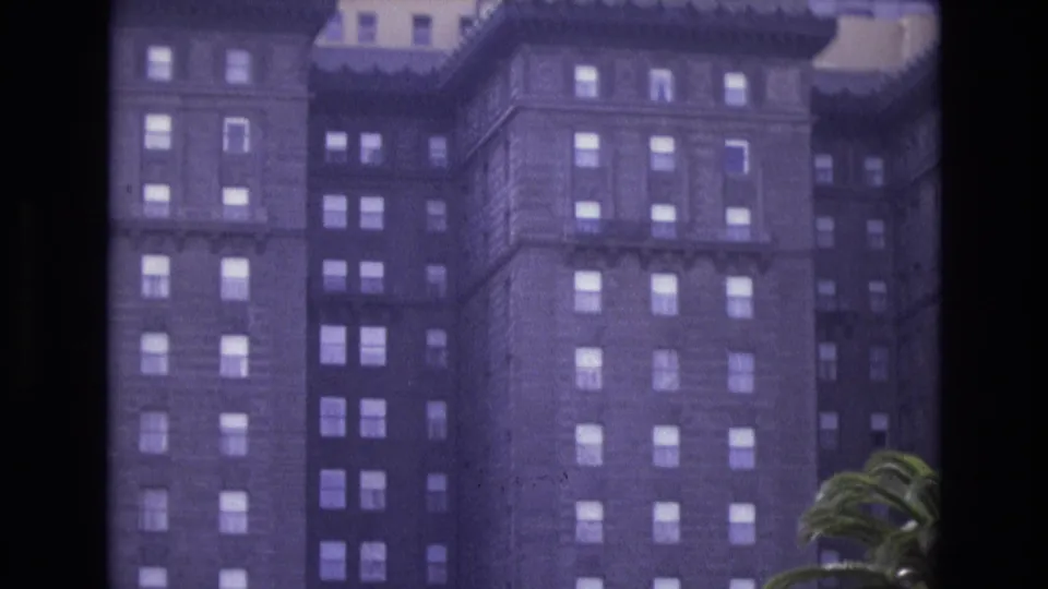 1973 San Francisco 8mm frame of a historic hotel exterior with repeated window rows, decorative upper trim, and palm fronds entering from the right.