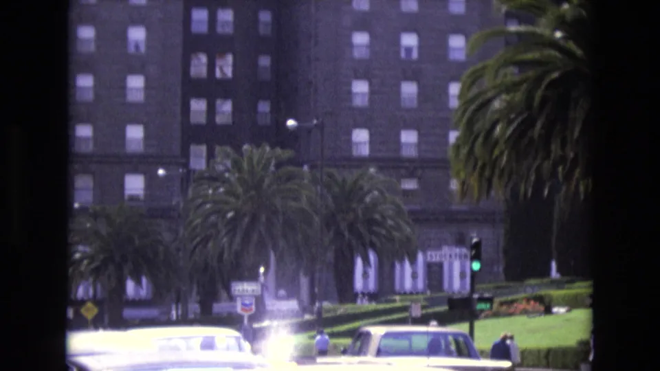 1973 San Francisco frame with bright period cars, pedestrians, palm trees, traffic lights, and a tall multi-story building in the background.