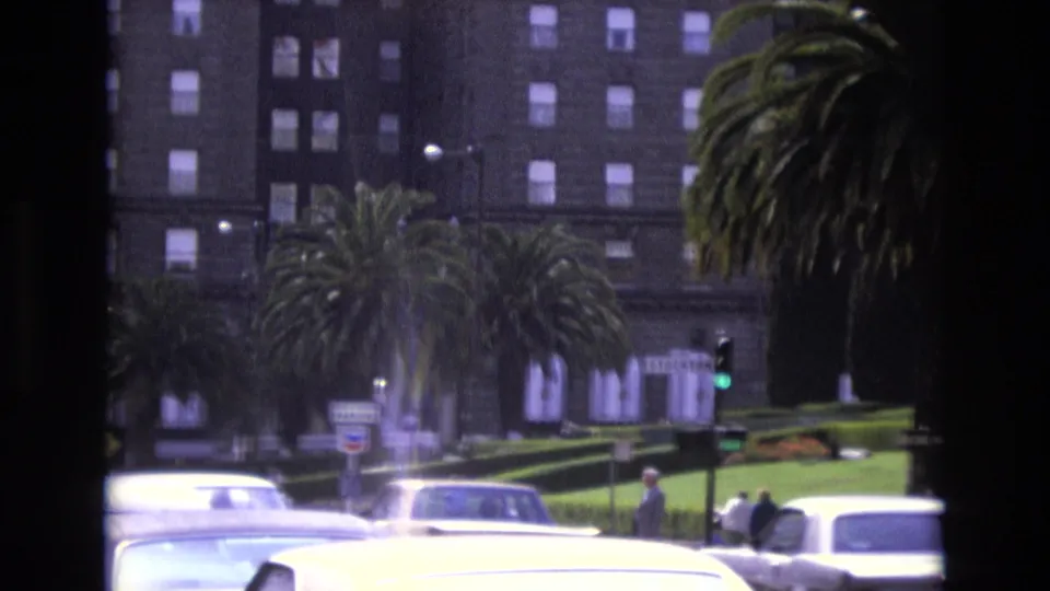 1973 San Francisco street view with overexposed passing cars, distant pedestrians, palm trees, a traffic signal, and a tall dark building behind them.