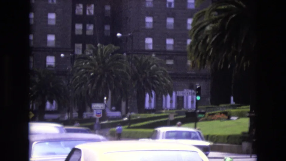 1973 San Francisco frame showing a busy road with classic cars, palm trees, street signage, pedestrians, and a tall windowed building in the background.