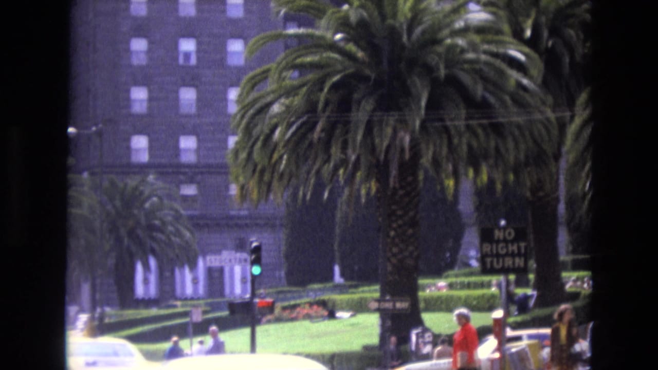 1973 San Francisco home-movie frame showing a palm-lined urban intersection with pedestrians, a green traffic light, cars, landscaped greenery, and a large building behind them.