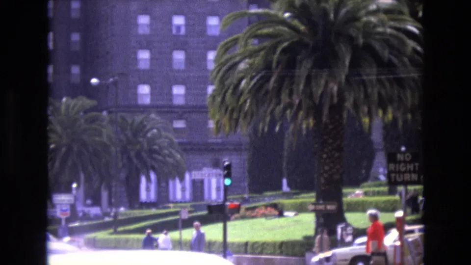 1973 San Francisco home-movie frame showing a tall palm tree before a large building, with pedestrians, a traffic light, cars, and street signs at the intersection.