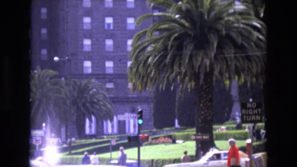 1973 San Francisco frame of a palm-lined intersection with pedestrians and cars moving through bright street glare, a green traffic light, street signage, and a multi-story building in the background.
