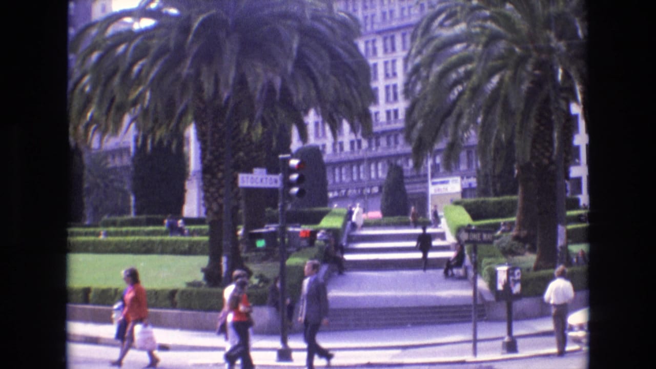 1973 San Francisco plaza-side street scene with pedestrians moving past palm trees, trimmed hedges, broad stairs, traffic signals, and visible street signage.