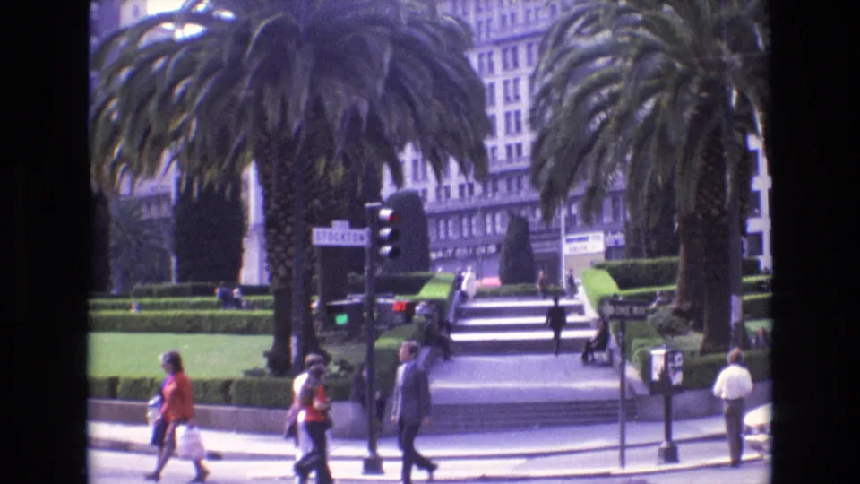 1973 San Francisco plaza-side street scene with pedestrians moving past palm trees, trimmed hedges, broad stairs, traffic signals, and visible street signage.