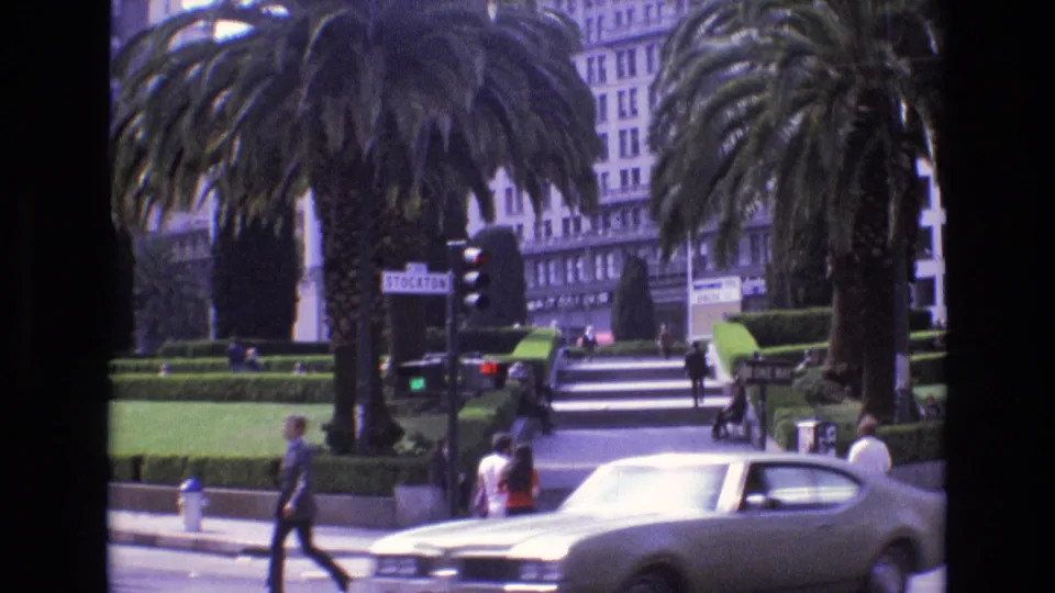 1973 San Francisco street footage showing a passing car in the foreground with pedestrians, palm trees, hedges, traffic signals, and stairs behind it.