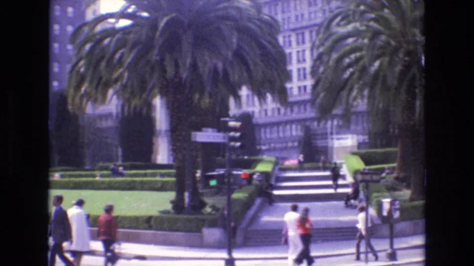 1973 San Francisco street scene with pedestrians crossing and walking near palm trees, traffic lights, manicured hedges, plaza stairs, and tall building facades.