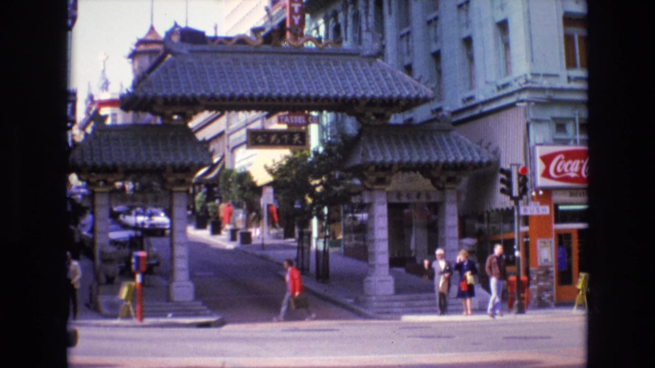 1973 8mm view of San Francisco Chinatown showing a Chinese-style gateway, pedestrians at the sidewalk, traffic lights, storefront signage, and a Coca-Cola sign.