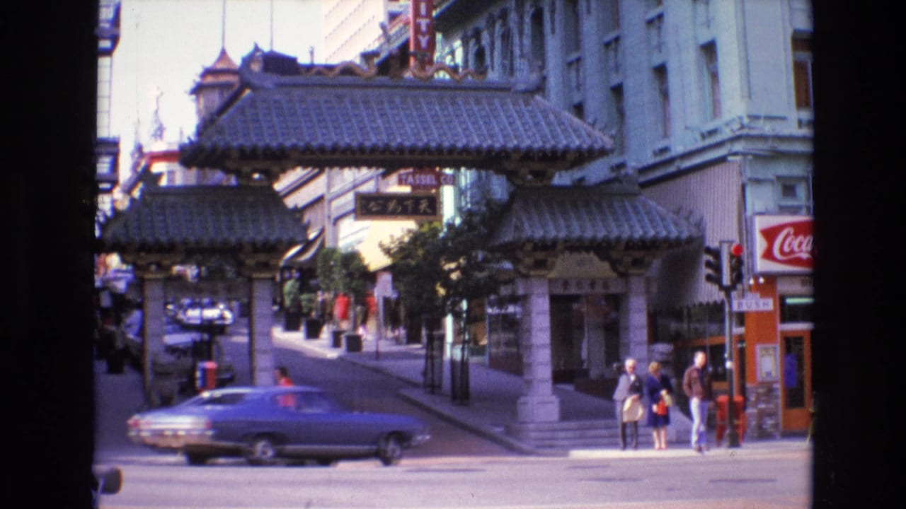 Car passing in front of the Chinatown gateway while pedestrians stand near storefronts.