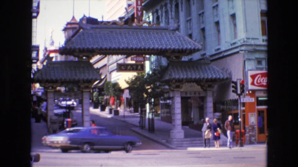 1973 San Francisco Chinatown street frame with a blue car crossing the foreground, the gateway behind it, pedestrians on the corner, and visible commercial signage.