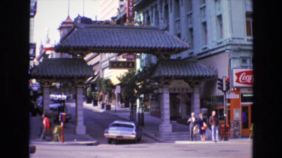1973 8mm frame of San Francisco Chinatown with the gateway centered, pedestrians near the curb, a light-colored car in the foreground, and storefront signage along the sidewalk.