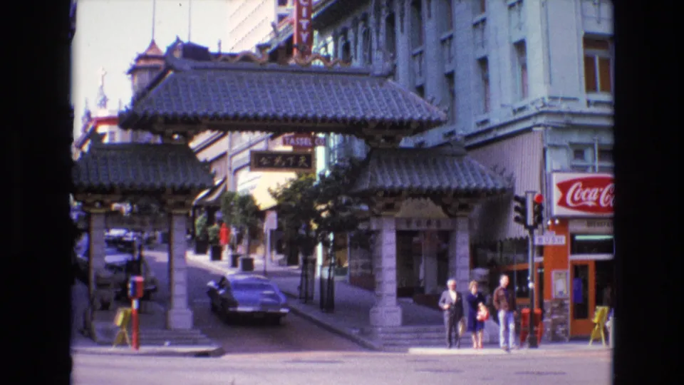 1973 San Francisco Chinatown view showing a dark car beneath or near the gateway, pedestrians on the right sidewalk, traffic lights, and commercial signs including Coca-Cola.