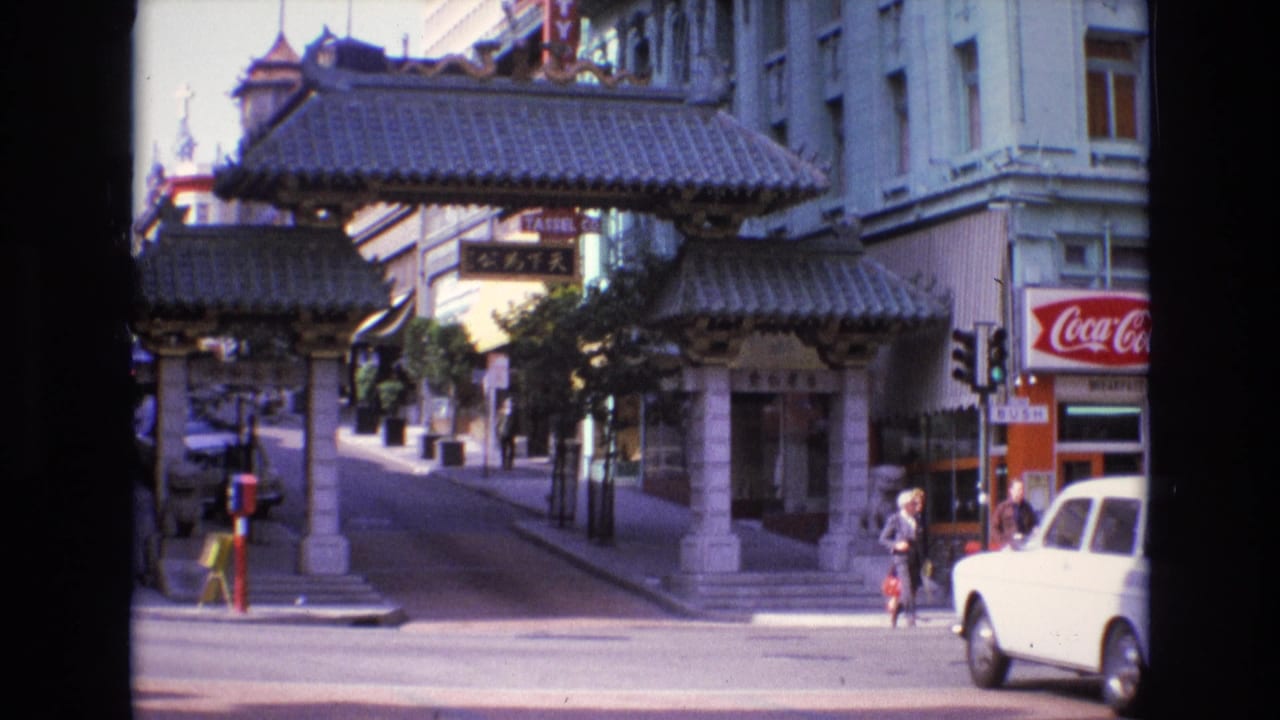White car enters the street scene beside the Chinatown gate and pedestrians.