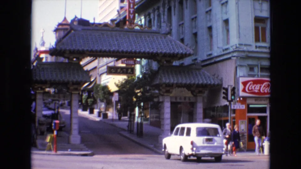 The white station wagon continues across the 1973 San Francisco Chinatown street scene, with the gate, stone columns, storefronts, and sidewalk pedestrians visible.