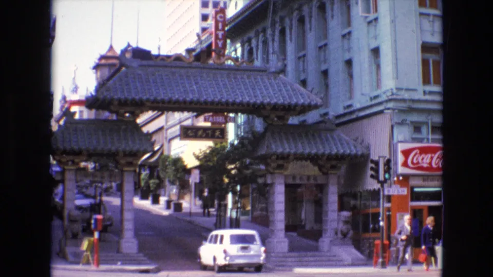 End frame of the San Francisco Chinatown gate scene, showing the white car farther along the street, pedestrians at the storefront corner, and 1970s urban signage.