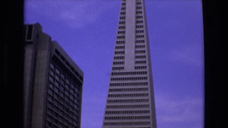 1973 San Francisco 8mm frame showing the Transamerica Pyramid from street level beside a neighboring high-rise, with grain and dark film border evident.