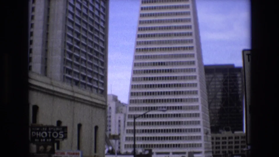 1973 San Francisco frame with the Transamerica Pyramid prominent at right, neighboring downtown buildings, a lamppost, and partial commercial signage near street level.