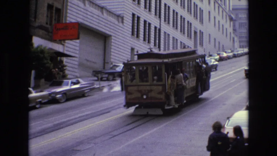 1973 San Francisco cable car scene with passengers riding on the side platform as the car moves along tracks past parked vehicles and tall downtown buildings.