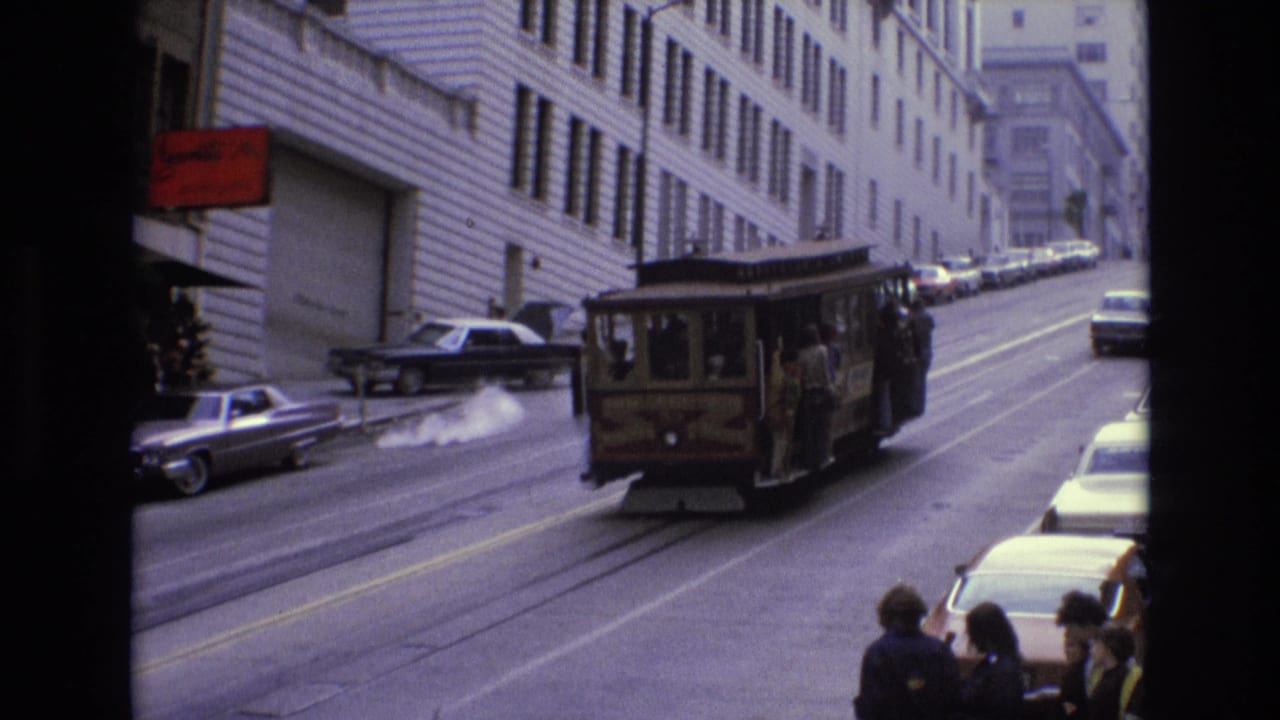 Cable car climbing a steep city street with cars, buildings, and pedestrians nearby.