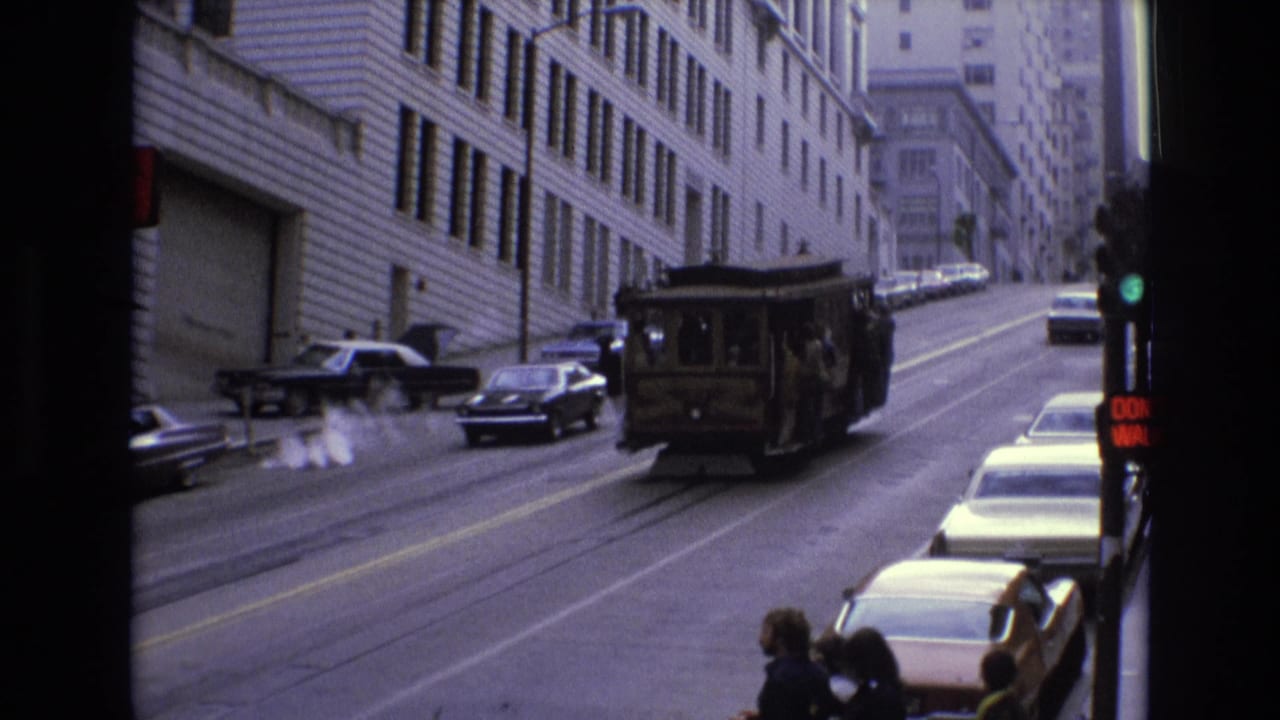 The cable car continues uphill on a San Francisco street, framed by mid-century cars, tall building facades, a traffic light, and people waiting near the curb.