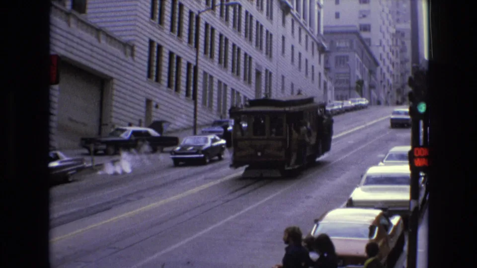 The cable car continues uphill on a San Francisco street, framed by mid-century cars, tall building facades, a traffic light, and people waiting near the curb.