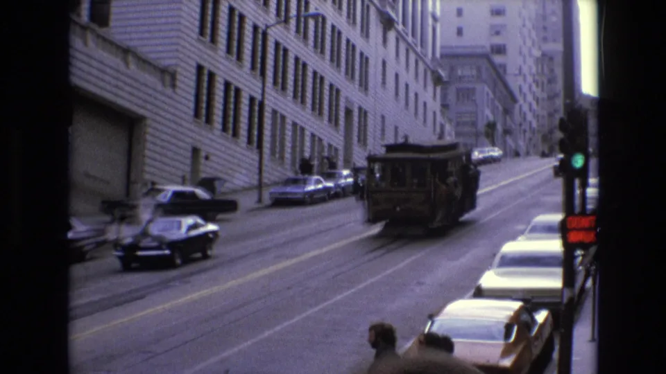 A cable car recedes up the steep San Francisco street while a green signal, rows of parked cars, building fronts, and corner pedestrians remain visible in the 8mm frame.