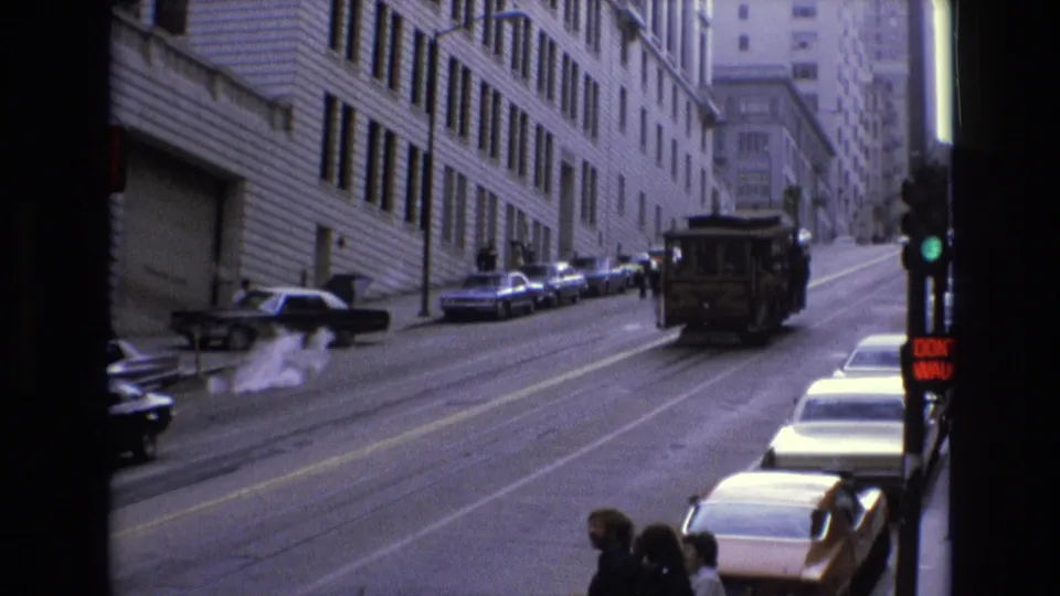Near the end of the shot, the cable car is farther uphill on the San Francisco street, with parked cars, pedestrians, a traffic light, and tall buildings still visible.