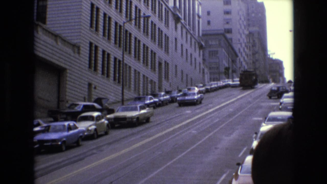 Steep city street lined with parked cars beside a large building, viewed through a dark film or vehicle frame.