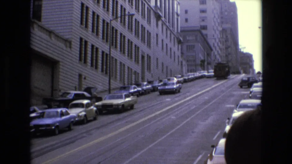 1973 San Francisco street view looking uphill past parked sedans and station wagons beside a large building with repeated windows.