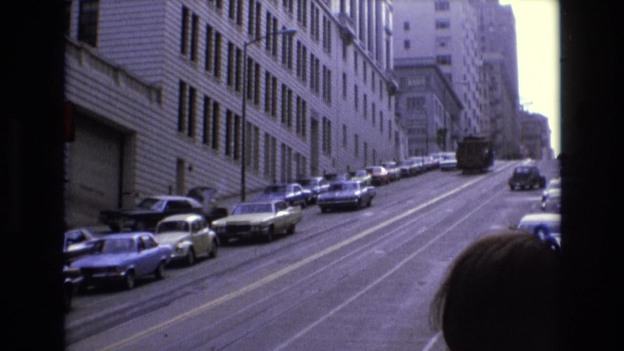 1973 San Francisco uphill street with rows of parked cars, a distant bus or truck, and a passenger silhouette partly visible in the foreground.