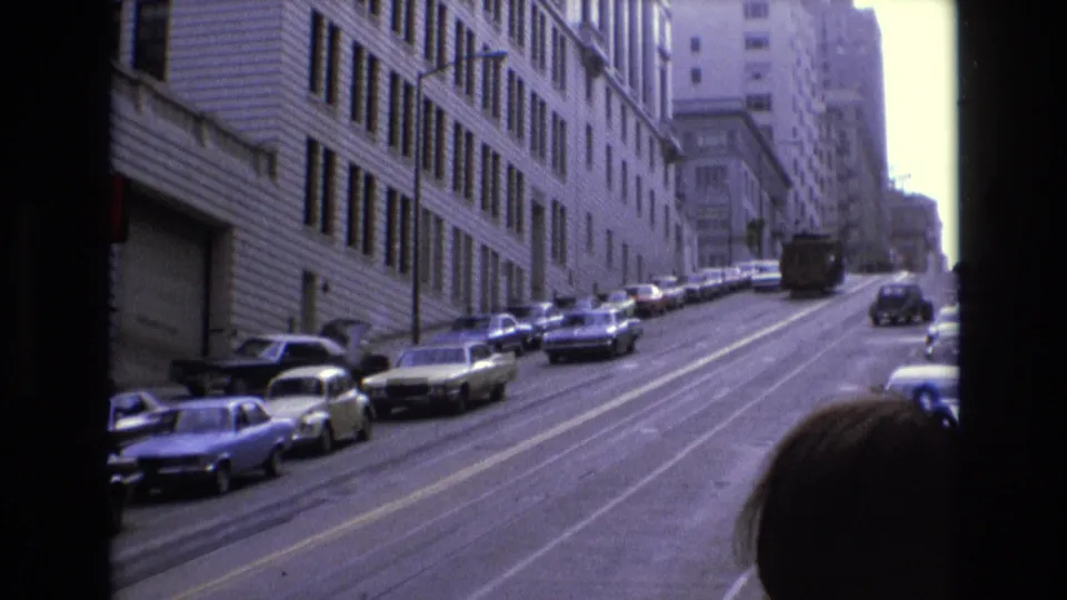 1973 San Francisco uphill street with rows of parked cars, a distant bus or truck, and a passenger silhouette partly visible in the foreground.