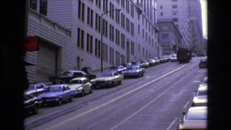 1973 San Francisco street frame showing parked vintage cars climbing a hill beside a large building, with a partial red sign visible at the left edge.