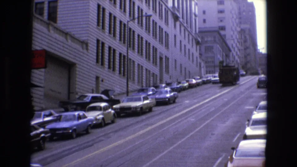 1973 San Francisco uphill street view with parked cars along both sides, a distant bus or truck, and a foreground passenger head silhouette at right.