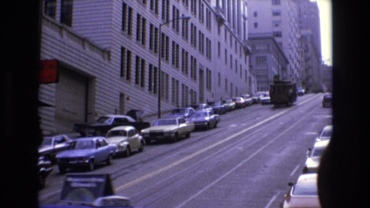 Steep city street with parked cars, rail tracks, and a distant cable car between tall buildings.