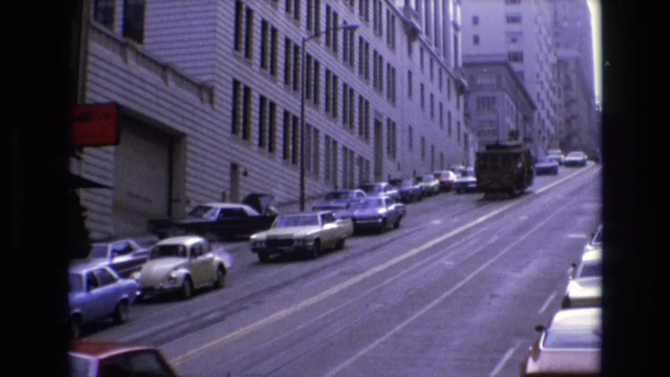 1973 San Francisco street view with a Volkswagen Beetle, several parked cars, rail tracks, and a cable car moving uphill between downtown buildings.
