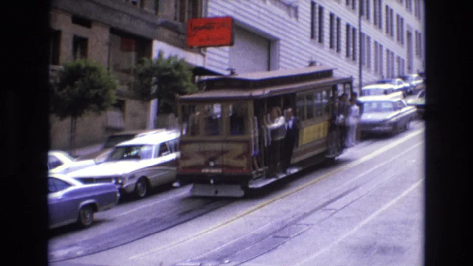 1973 San Francisco cable car passing close in the street, with visible passengers, parked cars, a red sign, and building facades along the hill.