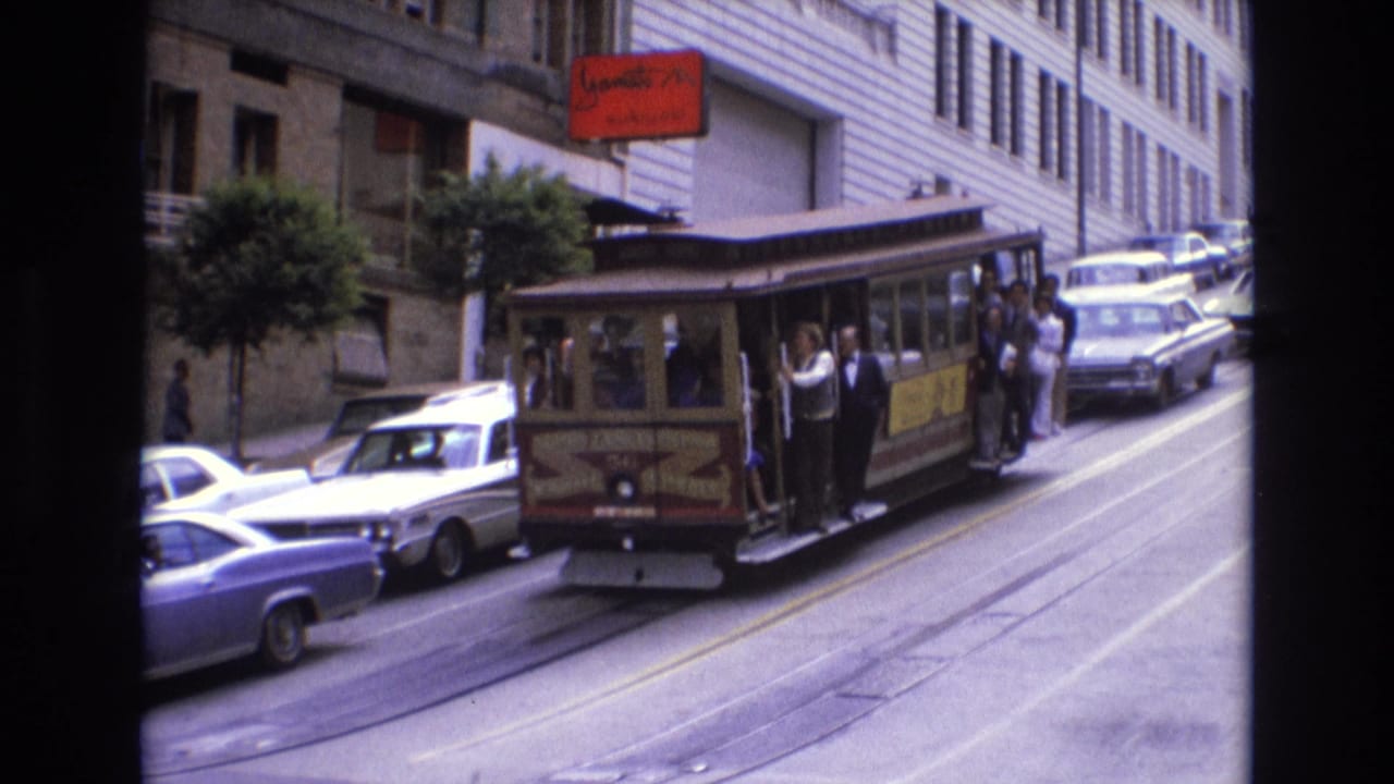 Cable car with passengers on a steep city street beside parked cars and tall buildings.